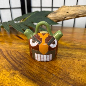 May include: A small, ceramic pitcher with a cartoonish face design. The pitcher features a brown body, orange beak, white teeth, and green handle and spouts. The face has angry eyes and a stern expression.