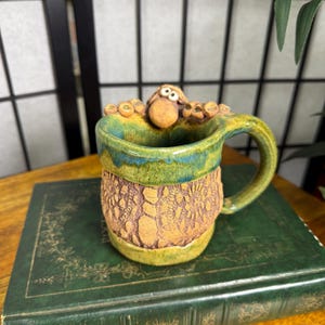 May include: A handmade ceramic mug with a whimsical sheep design. The mug features a textured brown body, a green and blue rim, and a curved handle. The sheep's head and ears peek over the rim. A unique, handcrafted piece.
