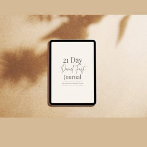 May include: A digital tablet with a black frame displays the text "21 Day Daniel Fast Journal" in a stylish font. The tablet is set against a neutral, textured background with subtle shadows, suggesting a natural setting. The journal is a 21-Day Prayer & Discipline Tracker.