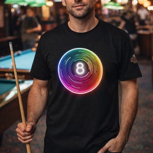 May include: Black t-shirt featuring a vibrant, multi-colored graphic of an eight-ball. The design has a neon-like glow. The person is holding a pool cue in a billiards room setting.