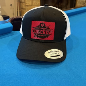 May include: Black and white trucker cap with a black peak and a red square patch. The patch displays "CUE CREW SUPPLY" with a pool ball and crossed cues graphic. The cap has a snapback closure.
