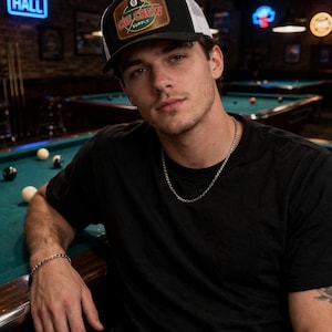 May include: A black and white trucker hat with a patch that reads "CUE CREW SUPPLY" and an 8-ball graphic. The hat is worn by a person in a black t-shirt, with a silver chain necklace and bracelet. The background shows a pool hall.
