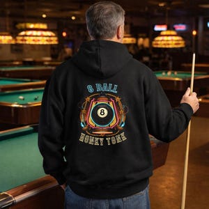 Puede incluir: Sudadera negra con un diseño de bola 8 dentro de una linterna de neón, con las palabras "8 BALL" y "HONKY TONK". La persona sostiene un taco de billar en un salón de billar.