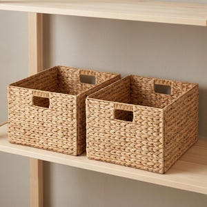 May include: Two woven storage baskets with rectangular cut-out handles. The baskets are made of a light brown, natural material and are sitting on a light-colored wooden shelf. The baskets are empty and ready for use.