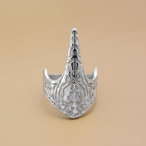 Puede incluir: Un anillo plateado con un diseño ornamentado. El anillo presenta un elemento central puntiagudo y patrones intrincados en su superficie. El diseño del anillo es simétrico y detallado, con una estética medieval o fantástica.