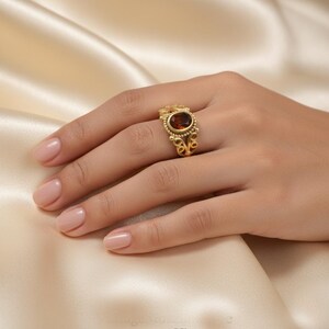 Könnte beinhalten: Ein goldener Ring mit einem ovalen, dunkelbernsteinfarbenen Stein. Der Ring hat ein verziertes Design mit kleinen, runden Akzenten und dekorativen Wirbeln. Der Ring befindet sich an einer Hand mit gepflegten Nägeln, vor einem cremefarbenen Stoff.