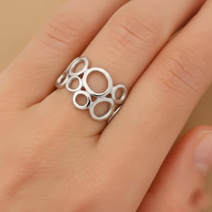 May include: A silver ring featuring an openwork design of interconnected circles of varying sizes. The ring is worn on a finger, showcasing its modern and abstract aesthetic. The ring's polished surface reflects light, highlighting its metallic sheen.