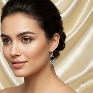 Może przedstawiać: Srebrne kolczyki wiszące z czarnym owalnym kamieniem i motywem liści. Kolczyki są prezentowane na osobie z ciemnymi włosami na neutralnym tle.