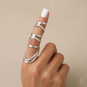 May include: A silver-colored ring set worn on a finger. The set includes multiple rings connected by bars, with a large, curved ring at the base. The rings have a polished, reflective surface.