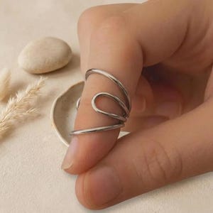 Könnte beinhalten: Ein silberfarbener Ring mit einem einzigartigen, fließenden Design. Der Ring umschließt den Finger mit einer Reihe von geschwungenen Linien und erzeugt einen abstrakten und modernen Look. Der Ring wird auf einem Finger mit neutralem Hintergrund gezeigt.
