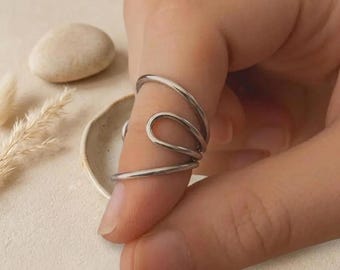 Puede incluir: Un anillo plateado con un diseño único y fluido. El anillo se envuelve alrededor del dedo con una serie de líneas curvas, creando un aspecto abstracto y moderno. El anillo se muestra en un dedo, con un fondo neutro.