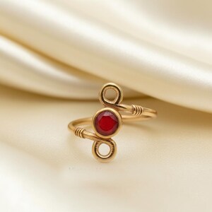Op de afbeelding: Een goudkleurige ring met een rode edelsteen in het midden. De ring heeft een uniek ontwerp met twee cirkelvormige lussen en een ronde, rode steen. De ring is geplaatst tegen een zachte, crèmekleurige achtergrond.