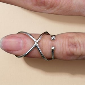 May include: A silver-colored finger ring with an X-shaped design and two small, round embellishments. The ring is worn on a finger, with the design wrapping around the finger.