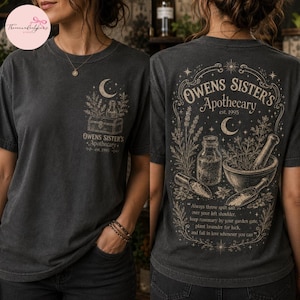 May include: A charcoal gray t-shirt featuring a graphic print. The front displays a small chest design of a chest, herbs, and a crescent moon with the text Owens Sister's Apothecary est. 1995. The back features a large, detailed illustration of a mortar and pestle, herbs, and a crescent moon, surrounded by ornate borders and the text Owens Sister's Apothecary est. 1995, along with a poetic quote about rosemary, lavender, and luck.