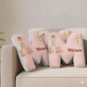 May include: Pink letter "M" shaped decorative pillows with a religious illustration of a woman, a cross, and the name "Miriam". The pillows have a light gray border and are decorated with pink roses.