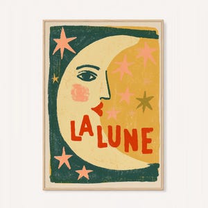 May include: A rectangular wall art print in a light wood frame featuring a stylized yellow crescent moon with a human face, red lips, and a pink cheek. The background is dark green with pink and gold stars and the red text LA LUNE.