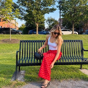 May include: A white racerback vest top paired with high-waisted red and pink checkered wide-leg trousers. The outfit is accessorised with a small black shoulder bag and black strappy heeled sandals. The look is completed with dark sunglasses.