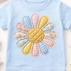 May include: A light blue t-shirt featuring a large, appliqued flower design. The flower has a yellow quilted center and alternating petals in pink gingham, floral, and blue gingham patterns. The shirt is likely for a child.