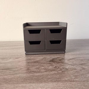 May include: A small, dark gray four-drawer storage unit. The drawers are rectangular with a recessed handle design. The unit is sitting on a wooden surface, with a neutral-colored wall in the background. The unit is designed for small items.