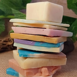 Puede incluir: Una pila de jabones hechos a mano en varios colores, incluyendo rosa, azul, amarillo y morado. Los jabones están apilados uno encima del otro, con un jabón cuadrado en la parte superior. El fondo es una tela de yute natural.