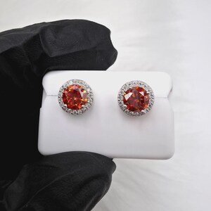 May include: A pair of stud earrings featuring a large, round, orange-red gemstone surrounded by a halo of smaller, clear stones. The earrings have a silver-colored setting and are displayed on a white surface.