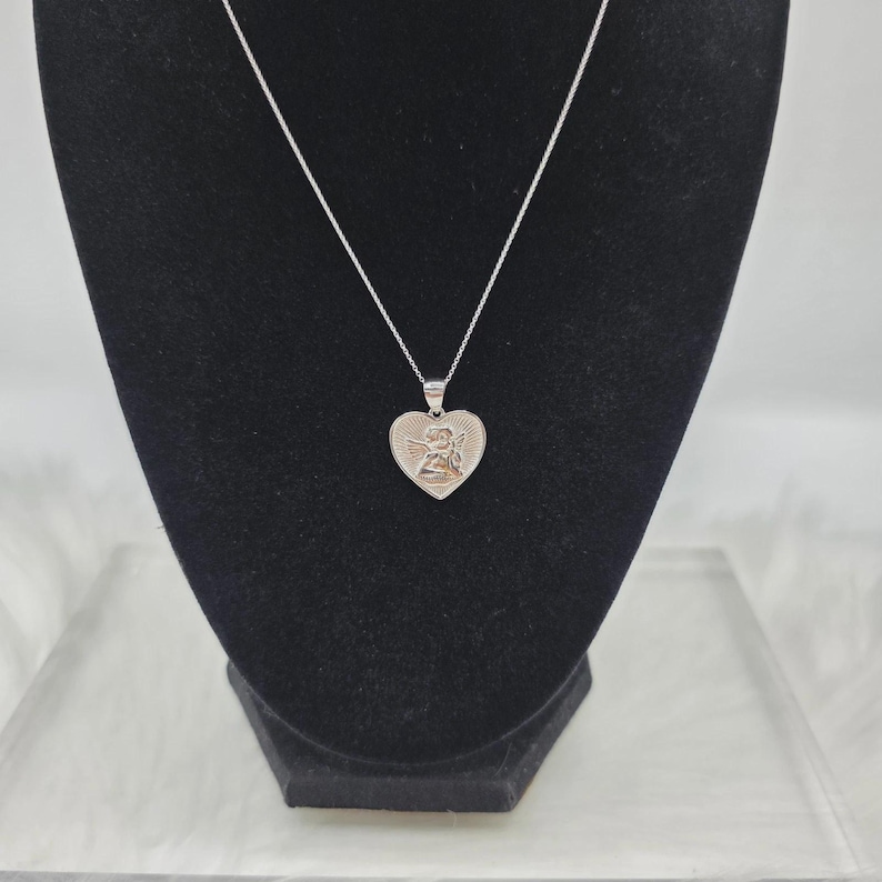 May include: A silver-toned heart pendant necklace. The heart-shaped pendant features an angel design and is suspended from a delicate chain. The necklace is displayed on a black velvet bust, against a white background.
