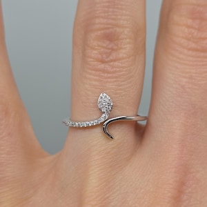May include: A silver ring in the form of a snake, featuring a head and tail design. The snake's head is adorned with small, clear gemstones, and the body is lined with similar gems. The ring is worn on a finger, showcasing a unique jewellery piece.