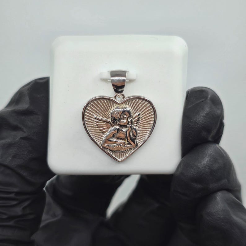 May include: Silver-toned heart pendant featuring a detailed cherub design. The pendant has a textured, sunburst-like background and a bail for a chain. The cherub is depicted with wings, adding a touch of elegance to this jewelry piece.