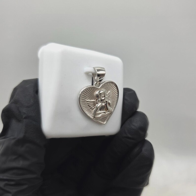 May include: Silver-toned pendant featuring a heart-shaped design with an angel figure in the center. The pendant has a bail for attaching to a chain. The angel is depicted with wings and a bow and arrow. The pendant is displayed on a white surface.