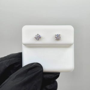 May include: A pair of sparkling, round-cut diamond stud earrings. The earrings are set on a white display card, held by a gloved hand. The diamonds are clear and reflect light, showcasing their brilliance. A classic jewelry item.