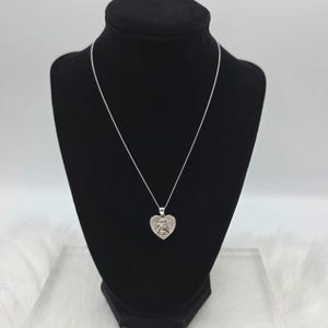 May include: A silver-toned necklace with a heart-shaped pendant. The pendant features a detailed design of a figure. The necklace is displayed on a black mannequin.