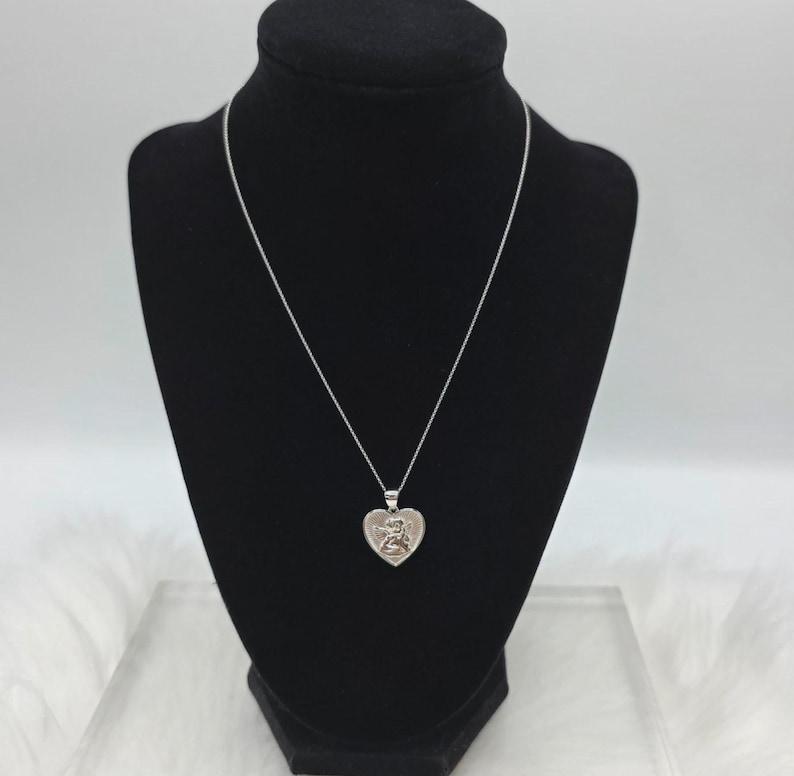 May include: A silver-toned necklace with a heart-shaped pendant. The pendant features a detailed design of a figure. The necklace is displayed on a black mannequin.