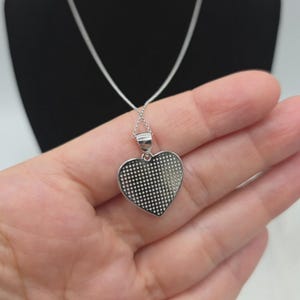 May include: A silver heart pendant necklace. The heart-shaped pendant has a textured surface with a dotted pattern. The necklace chain is also silver and delicate. The pendant is held in a hand.
