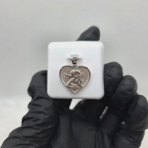 May include: Silver-toned heart pendant featuring an angel design. The pendant has a detailed relief of an angel within a heart shape, with sunburst rays emanating from behind. The pendant is displayed on a white surface.