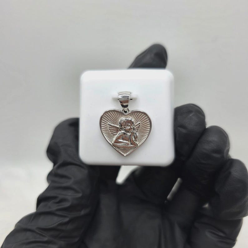May include: Silver-toned heart pendant featuring an angel design. The pendant has a detailed relief of an angel within a heart shape, with sunburst rays emanating from behind. The pendant is displayed on a white surface.