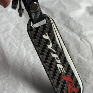May include: A black and grey carbon fibre keyring with the text "TYPE R" in white and red. The keyring has a silver metal ring and a black leather strap.