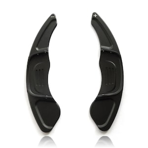 May include: A pair of matte black steering wheel paddle shifters with a curved, elongated design. The left paddle features a white minus sign symbol, while the right paddle features a white plus sign symbol. Both include textured grip sections.