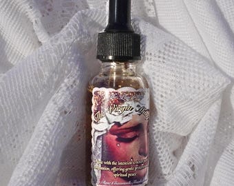May include: A small glass bottle with a black dropper top, filled with a light brown liquid. The label features an illustration of a face with closed eyes and the text "The Virgin Mary". The bottle rests on a white, textured fabric.