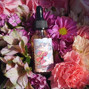 May include: A clear glass bottle with a black dropper is centered in a bouquet of pink, purple, and white flowers. The bottle has a label with a floral design and text. The flowers include roses, carnations, and chrysanthemums.