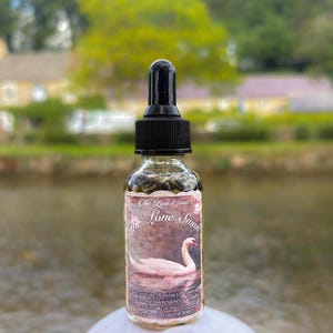 May include: A clear glass dropper bottle with a black cap and a label featuring a white swan on water. The label text reads The Lone Swan in a decorative script font. The bottle contains a dark liquid with visible botanical elements inside.