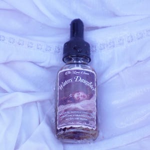 May include: A clear glass bottle with a black dropper top, labelled "Waters Daughter" with a vintage illustration. The bottle contains a liquid with visible herbs and sits on white, textured fabric.