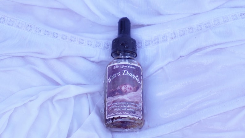 May include: A clear glass bottle with a black dropper top, labelled "Waters Daughter" with a vintage illustration. The bottle contains a liquid with visible herbs and sits on white, textured fabric.
