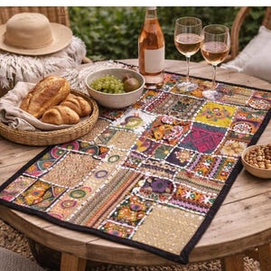 May include: A round wooden table set for a picnic with a colourful patchwork table runner. The table is set with wine glasses, a bottle of rosé, a basket of bread, a bowl of grapes, and a bowl of nuts. A straw hat rests on a chair.