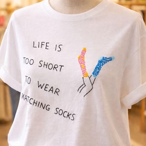 Puede incluir: Camiseta blanca con la frase "LIFE IS TOO SHORT TO WEAR MATCHING SOCKS". La camiseta presenta un diseño bordado de dos calcetines desparejados en rosa, amarillo y azul. Las mangas están dobladas.
