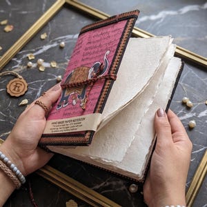 May include: A handmade paper notebook with a pink cover featuring a dark-coloured elephant illustration and black script text. The notebook has a black fabric border, a string closure, and thick, textured, off-white pages with deckled edges.