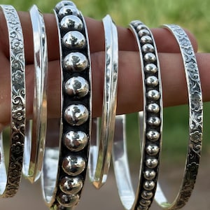 Peut inclure: Un ensemble de six bracelets rigides argentés aux designs variés. Deux bracelets présentent un motif floral en relief, deux sont lisses avec une arête centrale, et deux comportent une rangée de perles sphériques sur le centre de l'anneau.