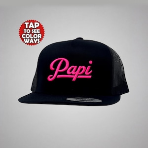 May include: Black trucker hat with a curved brim and mesh back. The word "Papi" is embroidered in bright pink script across the front. A red and white circular graphic reads "TAP TO SEE COLOR WAYS".