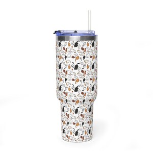 Op de afbeelding: Een witte beker met een blauw deksel en een doorzichtig rietje. De beker heeft een herhalend patroon van zwarte, bruine en oranje kattenillustraties. Ontworpen voor koude dranken.