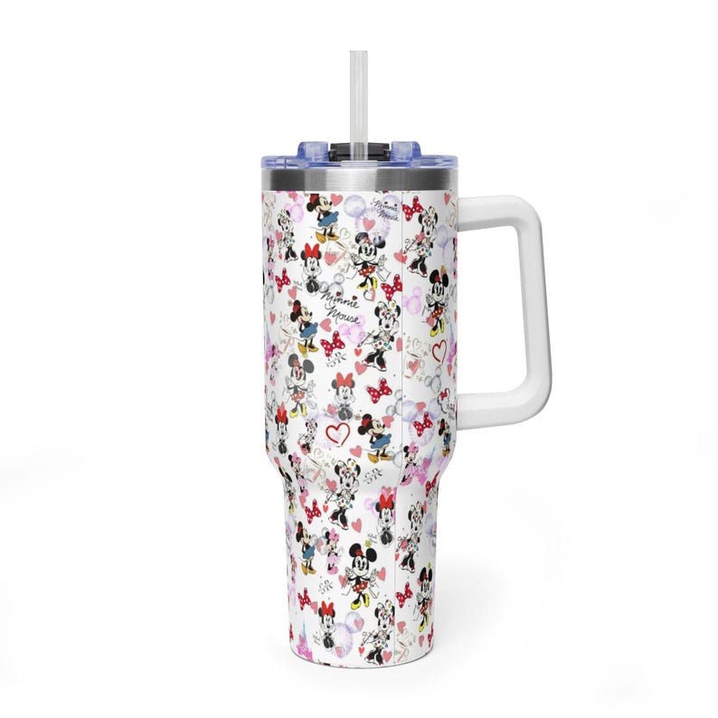 Puede incluir: Vaso blanco con asa y pajita, con un estampado repetido de ilustraciones de Minnie Mouse, corazones y lazos. El vaso tiene una tapa azul y un asa blanca. El texto "Minnie Mouse" es visible.