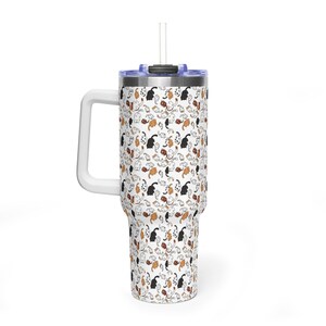 Op de afbeelding: Een witte beker met een handvat en een rietje. De beker is bedekt met een herhalend patroon van zwarte, oranje en witte kattenillustraties. Het deksel is blauw en het handvat is wit.